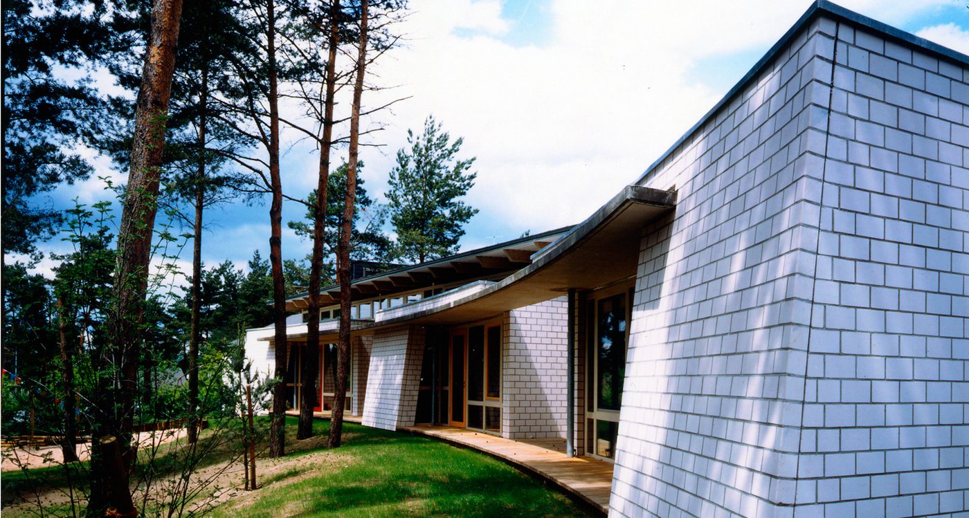 Kindergarten Edith Stein mit geschwungenem Dach und heller Schindelfassade zwischen Kiefern